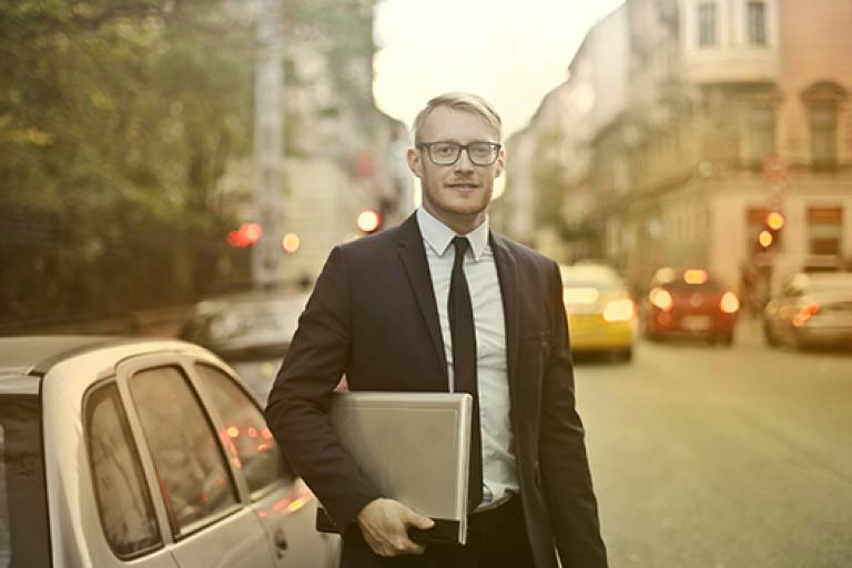 Man holding laptop standing next to a car on a busy city street